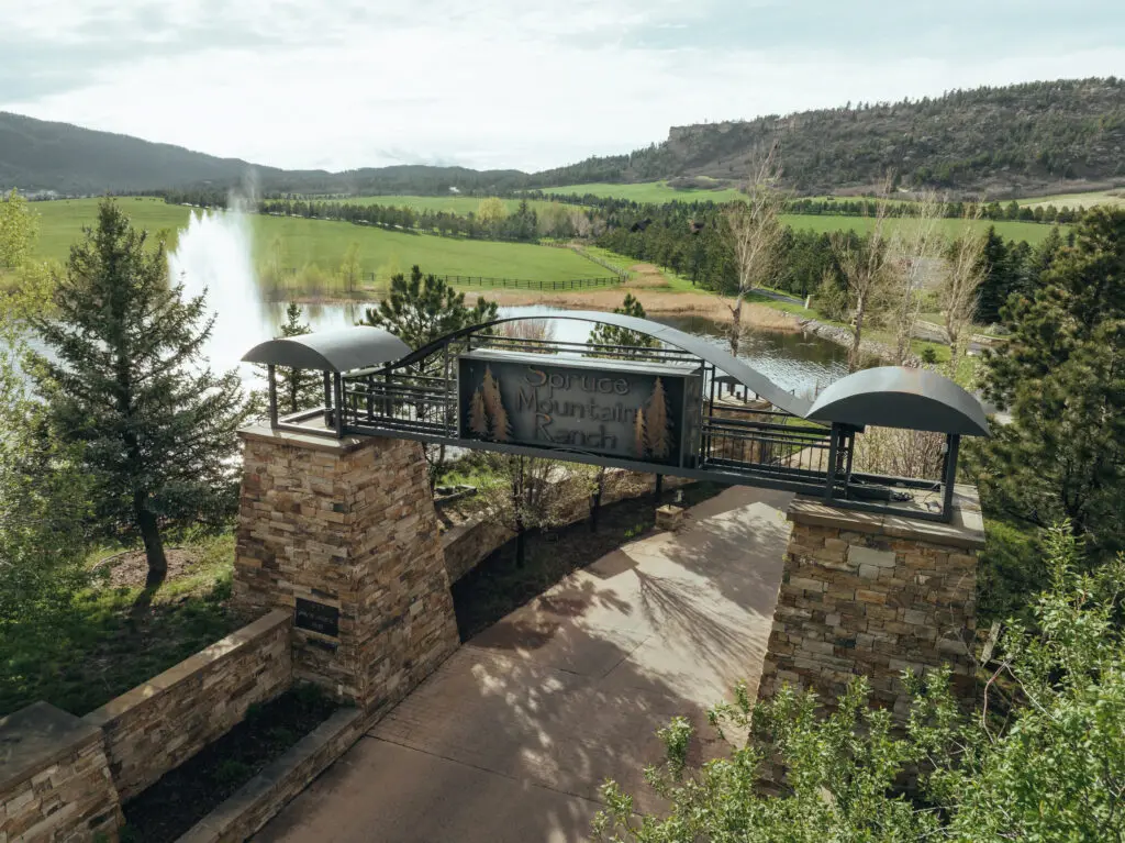  Spruce Mountain Ranch wedding venue entrance with rustic stone pillars, metal archway sign, scenic lake with fountain, green pastures and forested mountain ridges - premier wedding venues in Colorado