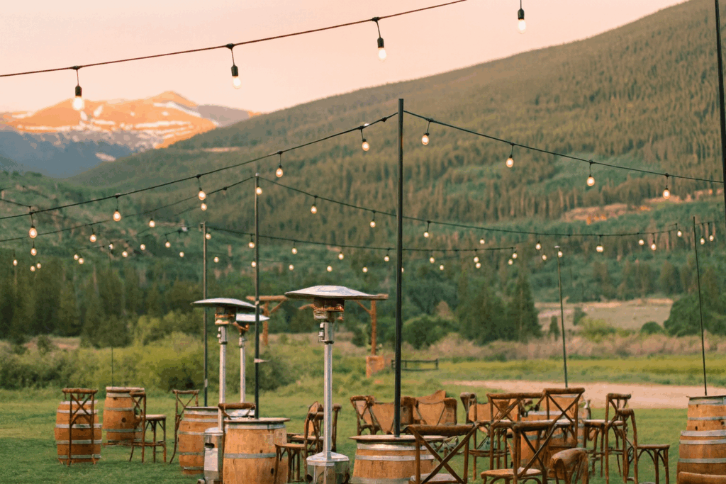 Rustic outdoor mountain wedding reception decorated with hanging string lights, wooden barrel tables, and patio heaters, overlooking sweeping alpine views and a golden sunset.