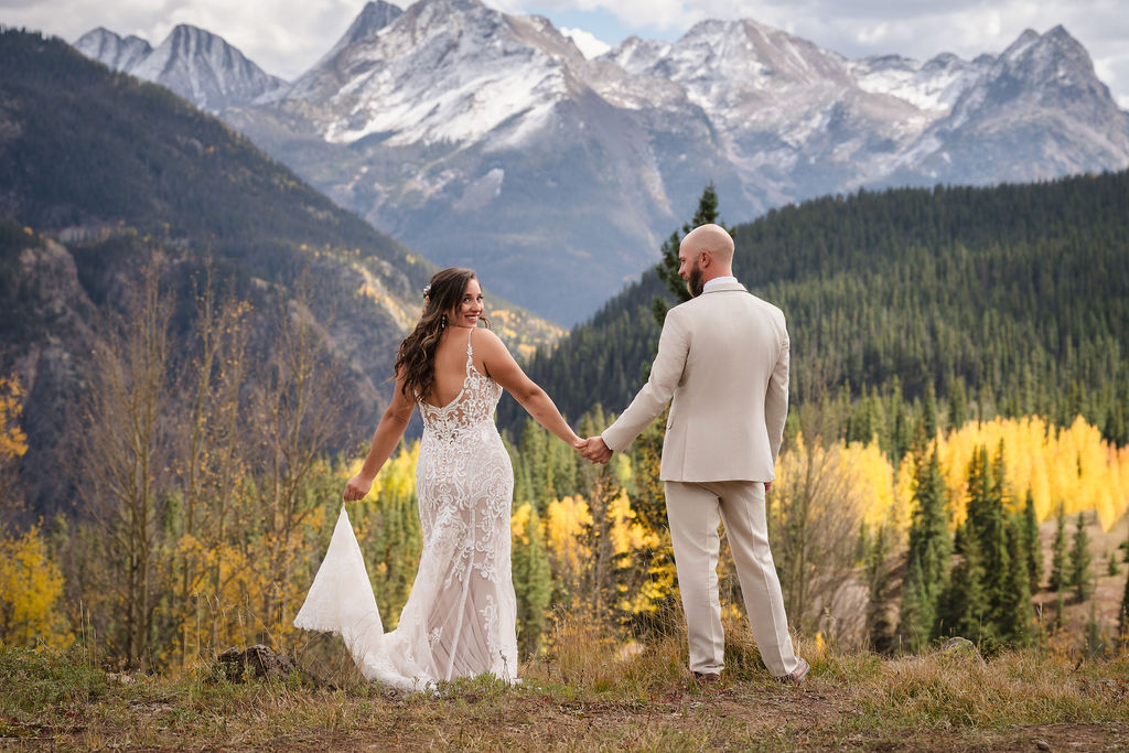 A horizontal wide shot of the couple holding hands and looking back toward the camera. The vastness of the Colorado wilderness and the snowy peaks create a cinematic setting for this wedding portrait.