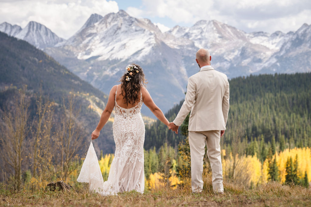 The bride and groom stand hand-in-hand, looking out toward a massive snow-dusted mountain range. A Silverton Colorado wedding photographer highlights the scale of the landscape as the bride holds her lace train.