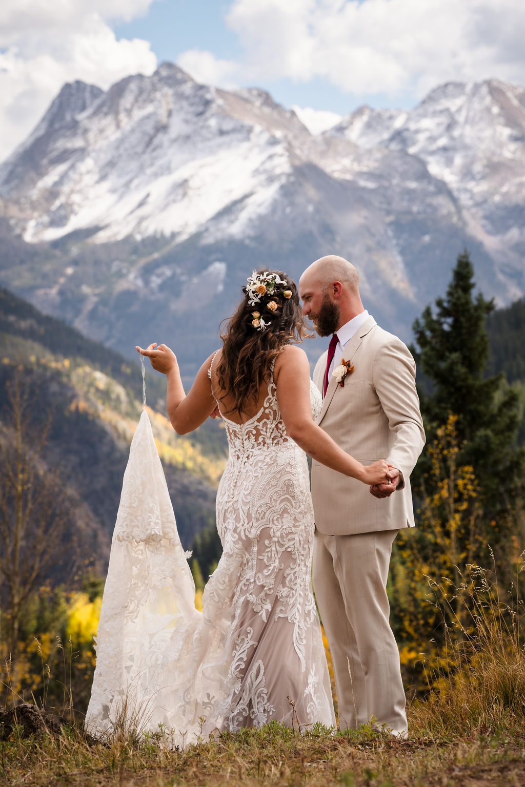 A side profile of the couple holding hands and smiling at one another. The bride's detailed lace gown and the groom's tan suit complement the natural, earthy tones of the high-country autumn.
