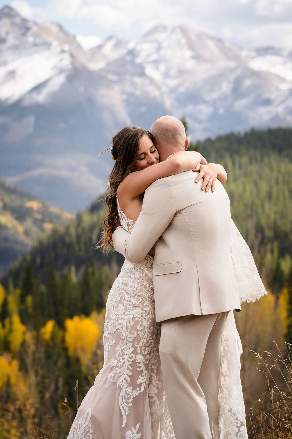 A tender moment where the bride rests her head on the groom's shoulder with her eyes closed, featuring the dramatic, jagged peaks of the Colorado Rockies in the blurred background.