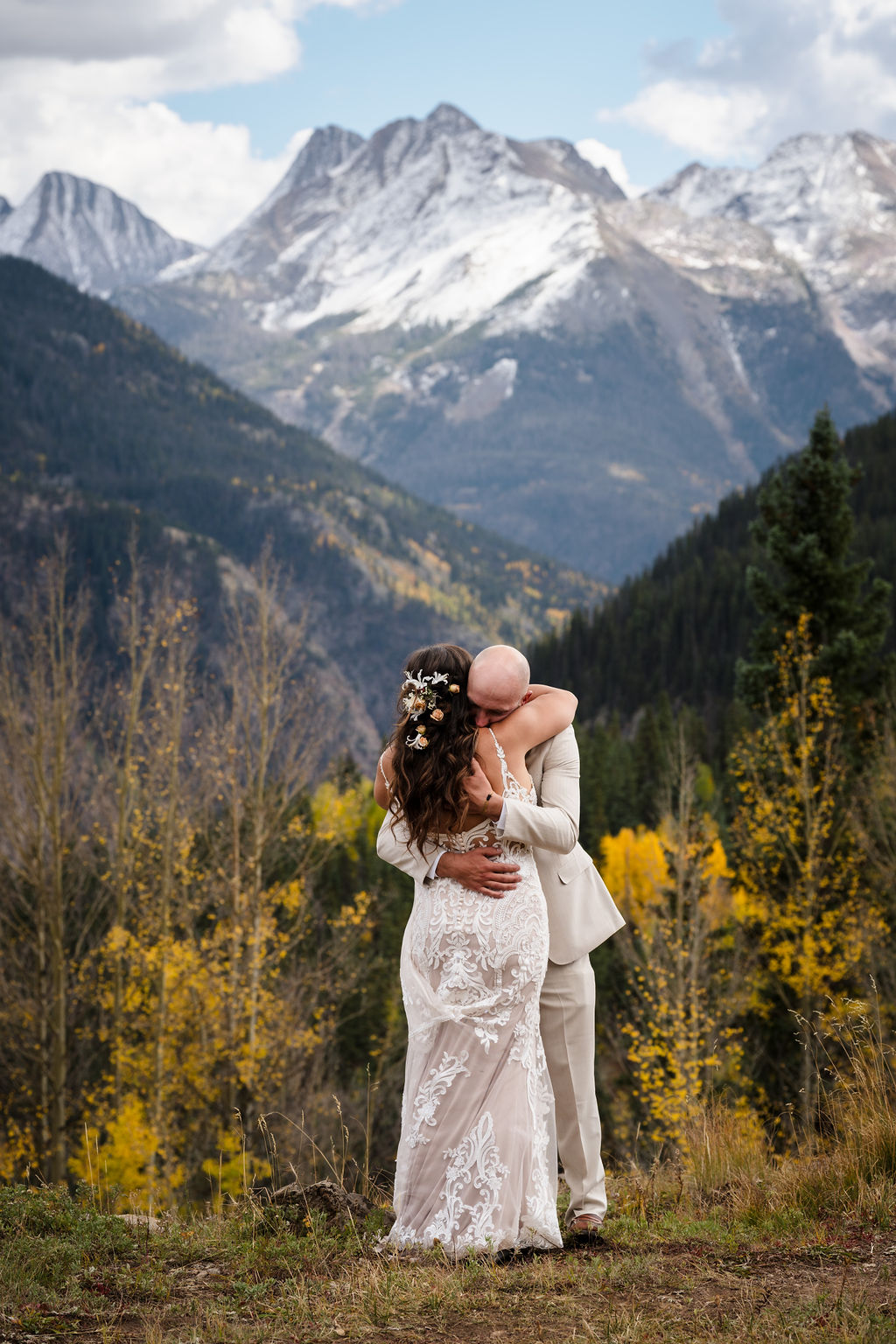 Wide shot of the couple embracing in a vast mountain valley. The bride’s intricate lace dress and floral hairpiece stand out against the deep greens and yellows of the Silverton landscape.