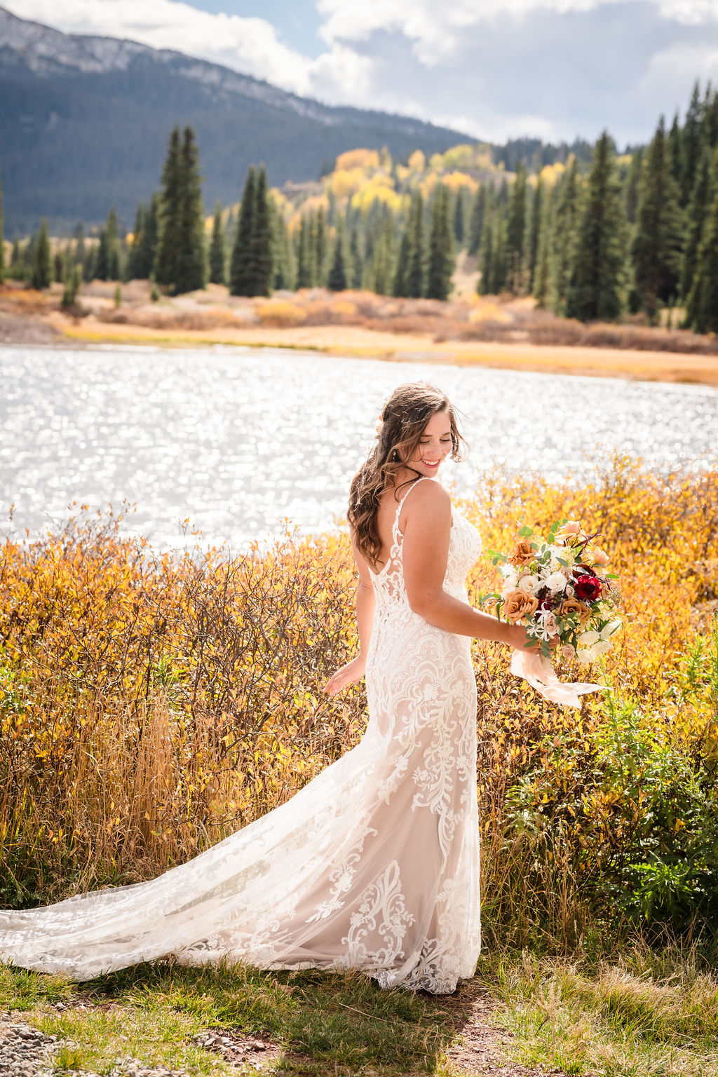 The bride poses by a sparkling alpine lake, holding a lush bouquet of fall-toned flowers with golden aspen trees and evergreen forests lining the shore.
