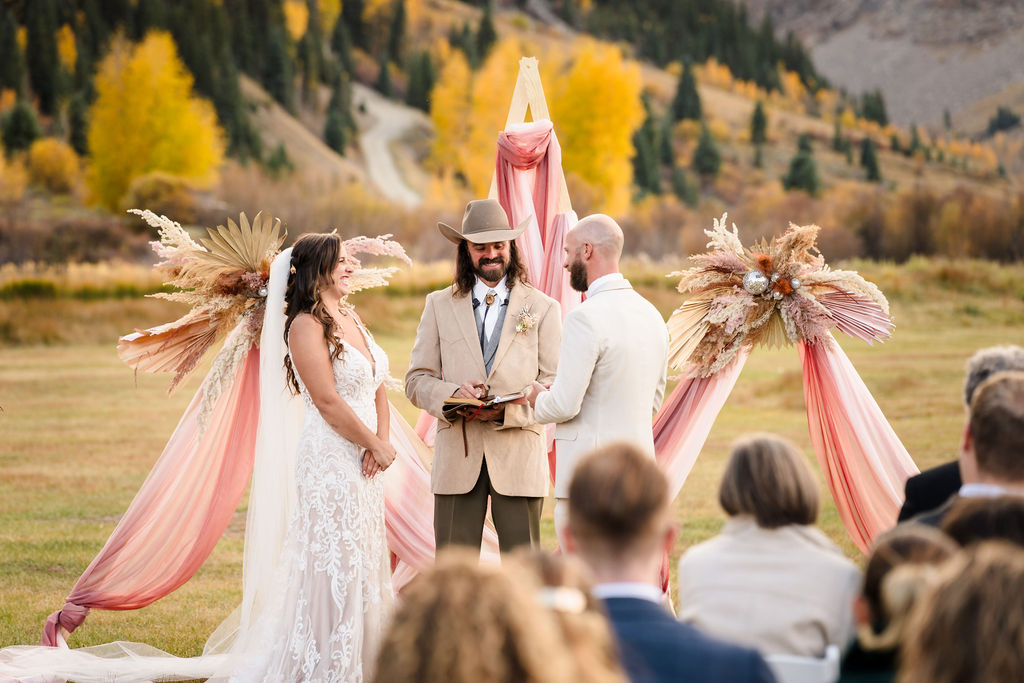 The bride and groom laugh together at the altar during their outdoor ceremony, framed by a boho-style wooden arch with pink draping, as captured by a Silverton Colorado wedding photographer.
