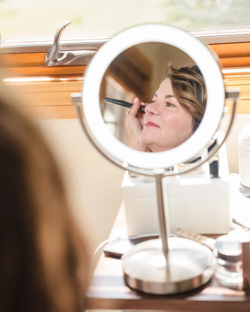 A close-up reflection in a lighted vanity mirror shows a woman applying makeup with a brush, preparing for a Chaparral Ranch Wedding.
