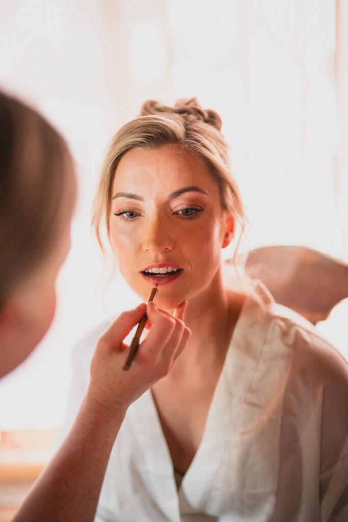 A tight close-up of a bride having her lip liner applied by a makeup artist. She is wearing a white silk robe, and the lighting is soft and warm.