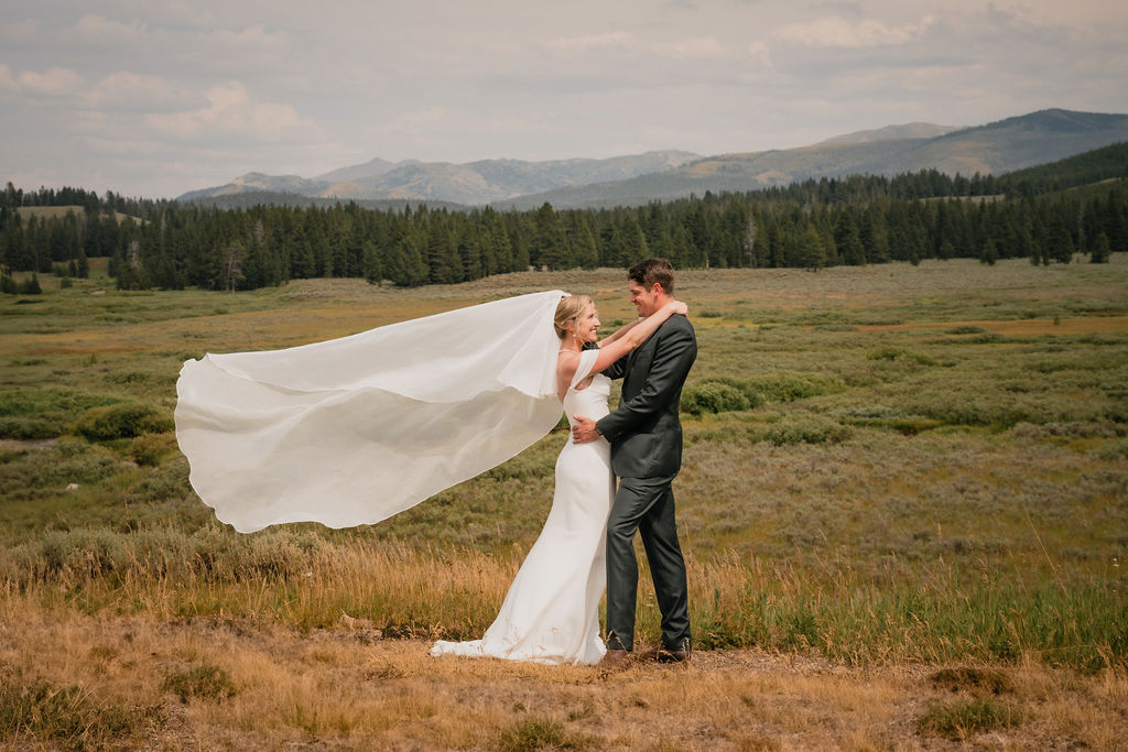 A wide shot of the couple embracing in a vast golden field. The bride's long white veil catches the wind, billowing out against the backdrop of distant evergreen forests and mountain peaks.