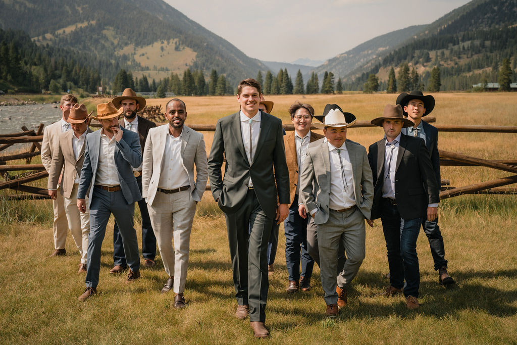 The groom leads a group of groomsmen, many wearing cowboy hats and suits, as they walk across a field with a river and mountains in the background.