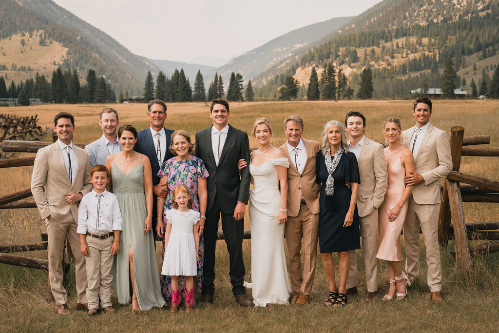 A formal wedding party and family portrait posed against a wooden ranch fence with the dramatic Montana mountain landscape in the distance.