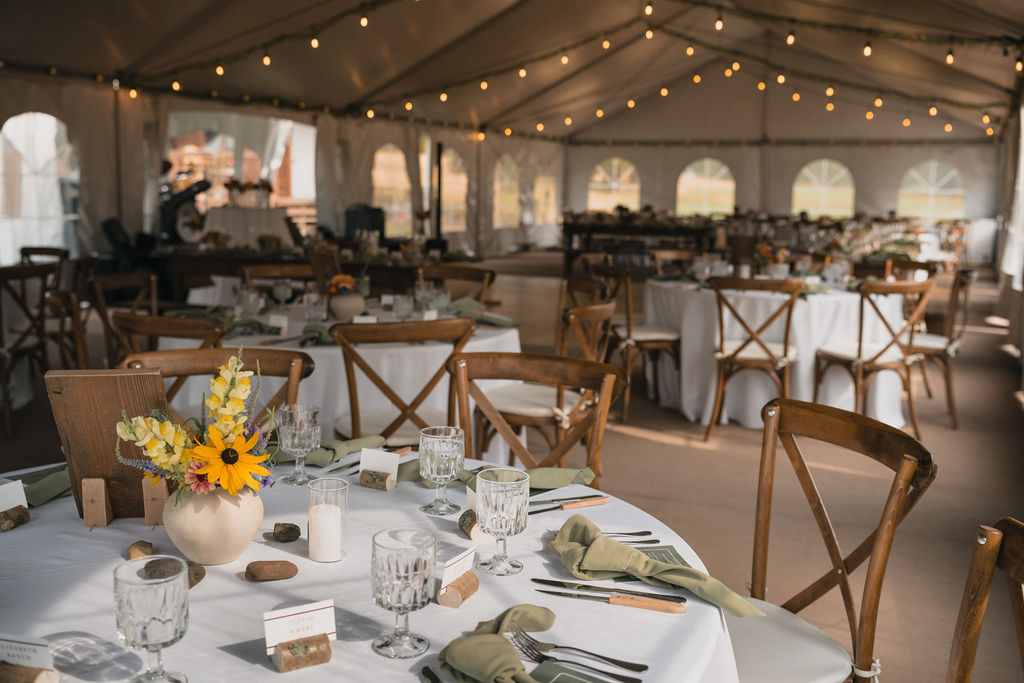 An interior view of a large white wedding tent set up for a Montana ranch wedding reception, featuring wooden cross-back chairs, white linens, and warm string lights overhead.