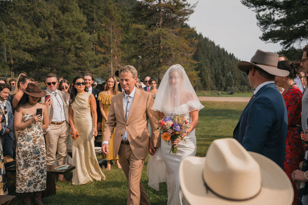 A bride in a white gown and long veil is escorted down a grassy aisle by a man in a tan suit at an outdoor Montana ranch wedding ceremony. Guests watch from the sides.