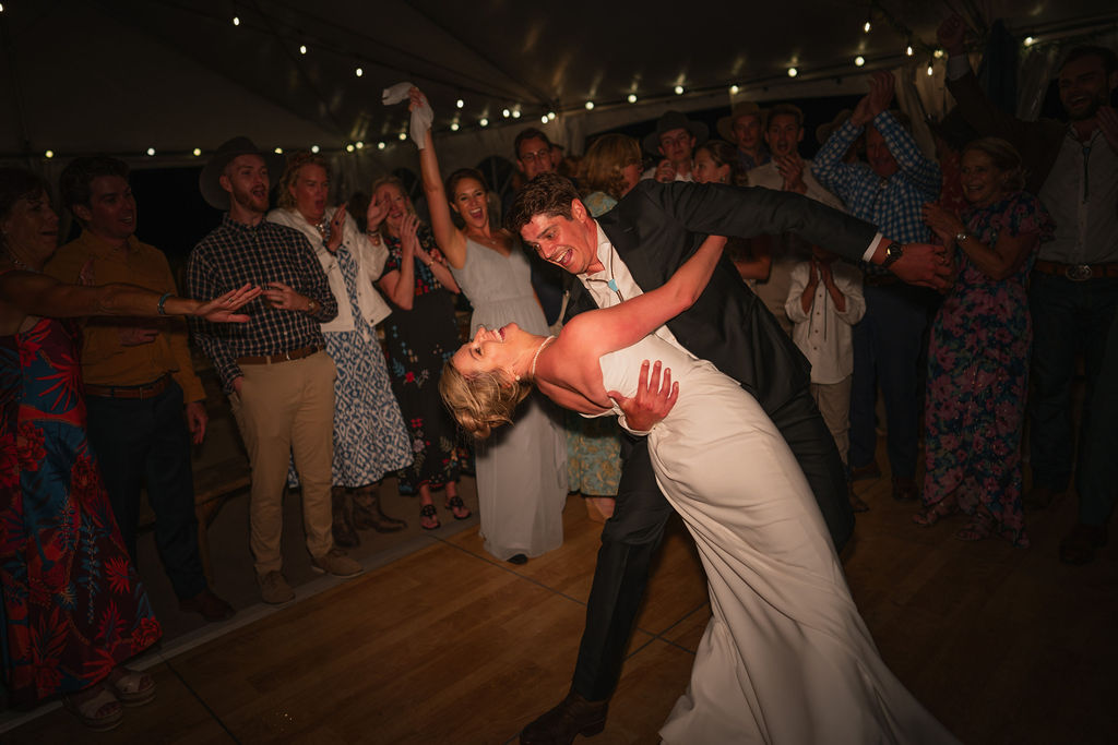 A high-energy photo of the groom dipping the bride on a wooden dance floor during their reception. Guests in the background are cheering and clapping under glowing string lights.