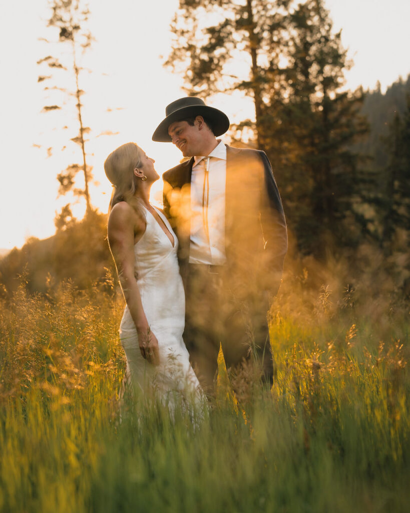 A romantic wide shot of the bride and groom standing in a tall grass field at sunset. The groom wears a cowboy hat and suit, and the bride wears a sleek white halter-neck gown. Golden light flares through the trees behind them.
