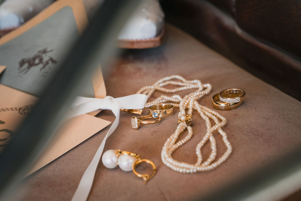A flat-lay detail shot of bridal accessories on a rustic leather surface, featuring a three-strand pearl necklace, gold rings including a large emerald-cut diamond and a blue sapphire ring, and large pearl drop earrings. A white ribbon and stationery with a cowboy illustration are visible in the background.