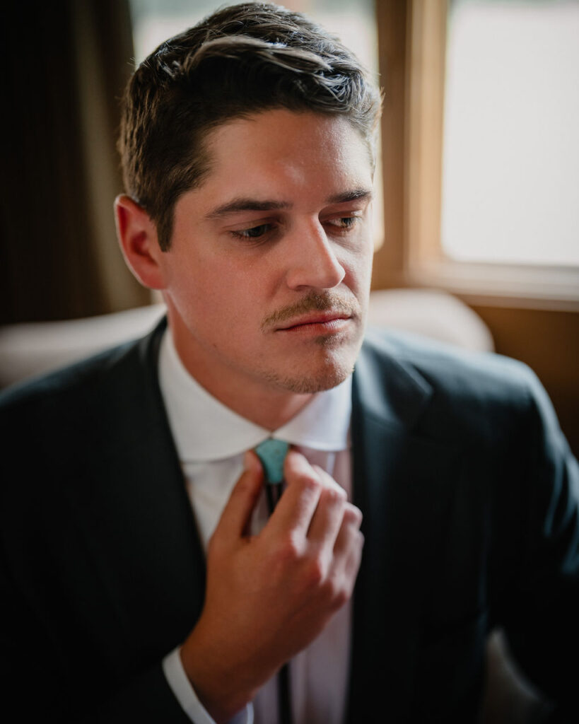 A close-up shot of the groom adjusting his turquoise bolo tie at the collar of his white dress shirt.