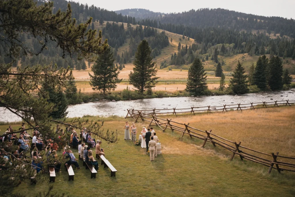 Outdoor wedding ceremony beside a river with mountain views, rustic wooden fencing, and guests seated on benches in Colorado.