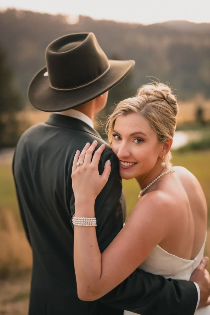 Bride smiling warmly over her groom’s shoulder during a golden-hour outdoor wedding, wearing elegant pearl jewelry while the groom stands in a dark suit and cowboy hat.