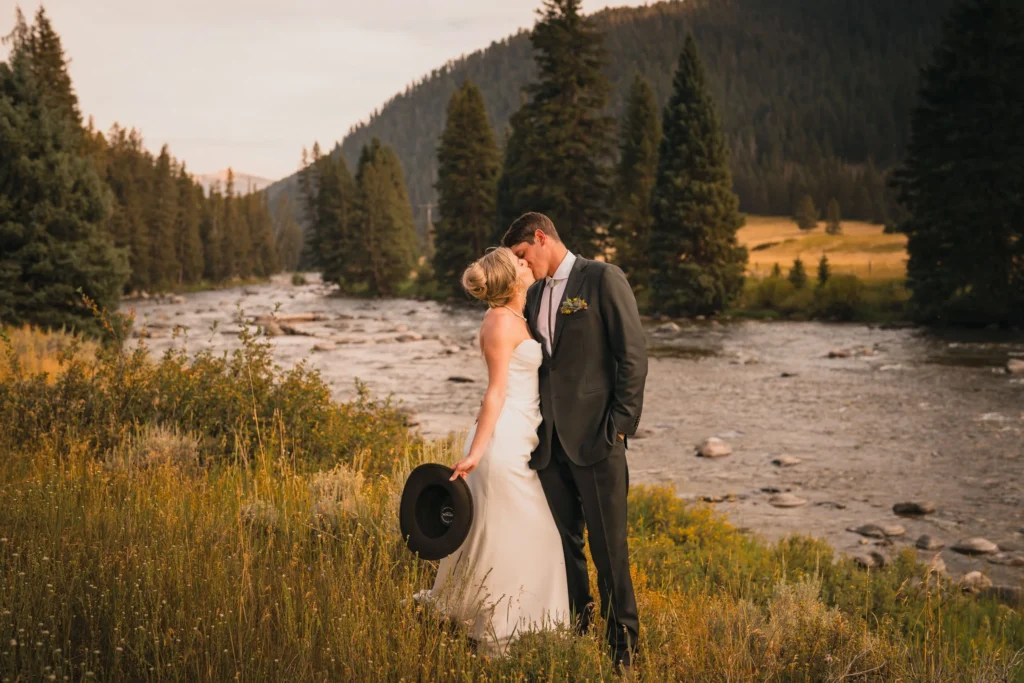 Romantic bride and groom kissing beside a flowing mountain river at sunset, surrounded by pine trees and golden meadow scenery.