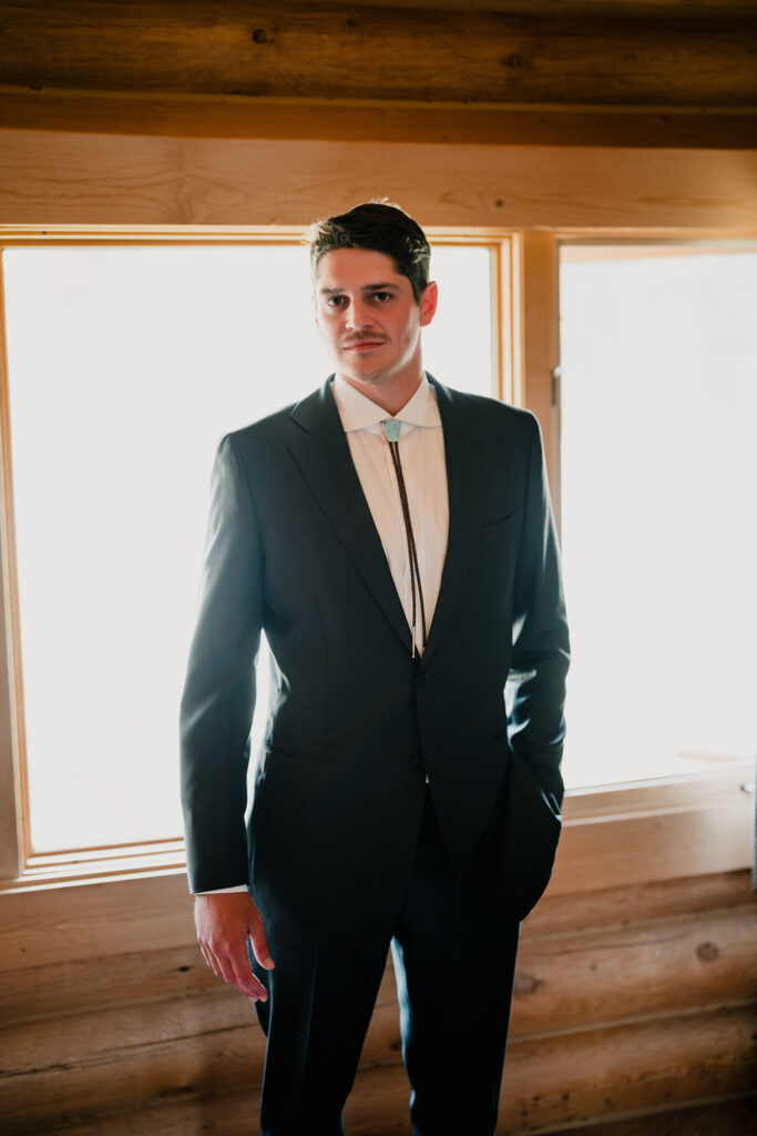 A full-body portrait of a groom in a dark suit and white shirt, wearing a turquoise bolo tie. He is standing in front of a bright window in a rustic log cabin.