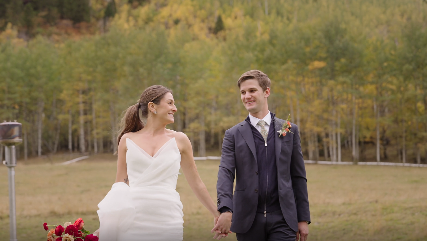 A bride and groom walk hand-in-hand through a grassy field during their Aspen Ranch Wedding, with a backdrop of golden autumnal trees and mountains.