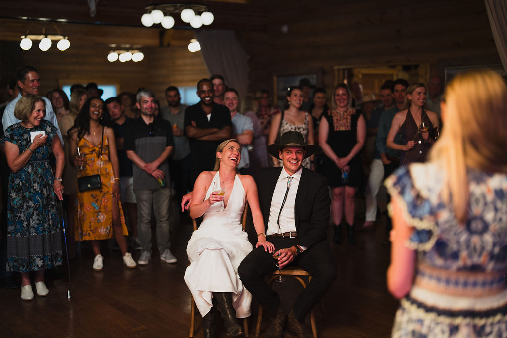 A bride and groom in Western-inspired wedding attire sit laughing while listening to a toast. They are surrounded by a crowd of smiling guests in a warm, rustic venue.