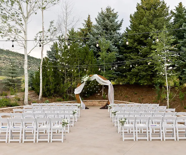 Outdoor wedding ceremony setup with white folding chairs, fabric-draped wooden arch, string lights, evergreen trees and mountain hillside