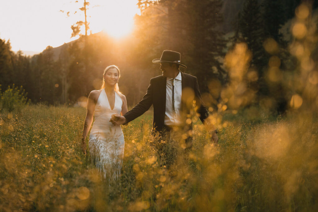Romantic sunset wedding portrait of bride in halter dress and groom in cowboy hat walking through golden wildflower meadow with warm bokeh and mountain backdrop