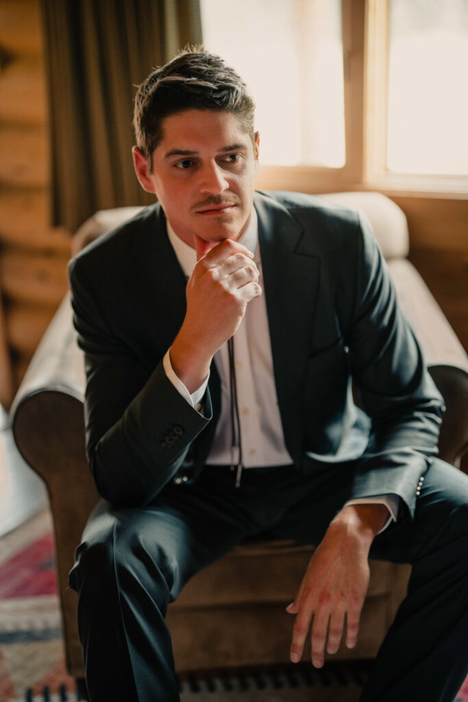 Groom in navy suit sitting pensively on ottoman in elegant hotel room with natural window light
