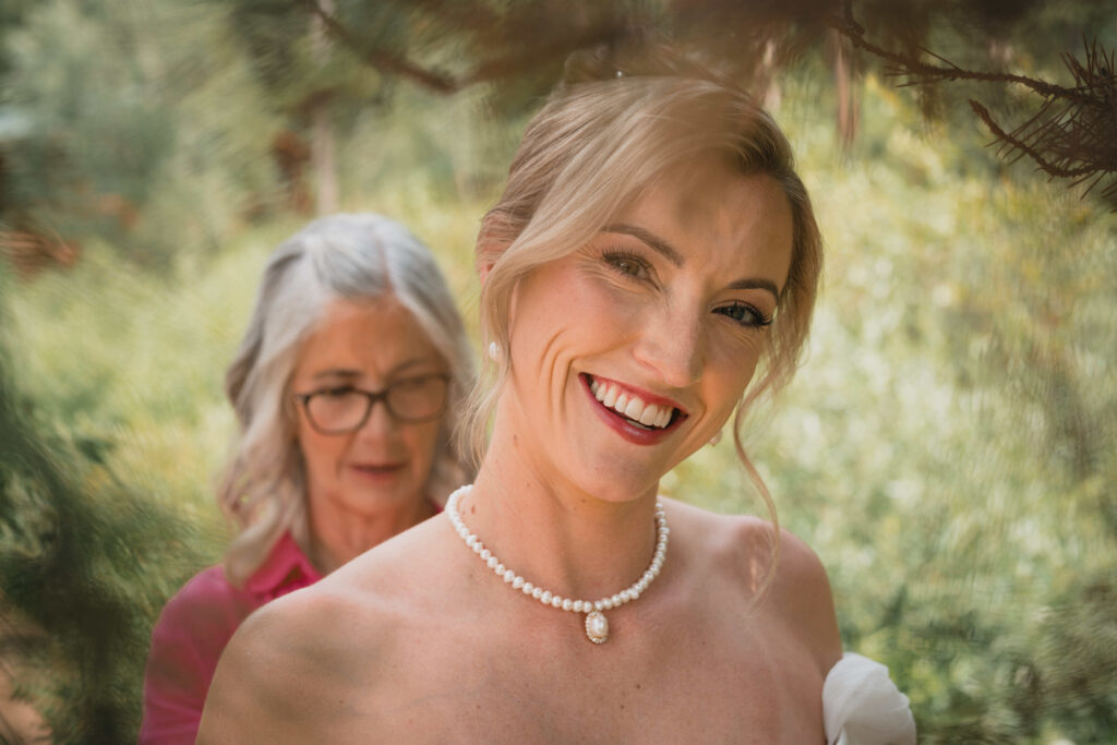 Radiant bride with upswept blonde hair wearing pearl necklace smiling at camera with mother behind in pine forest