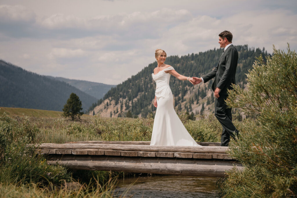 A bride and groom hold hands while standing on a rustic wooden bridge surrounded by mountains and greenery, celebrating their elegant wedding .