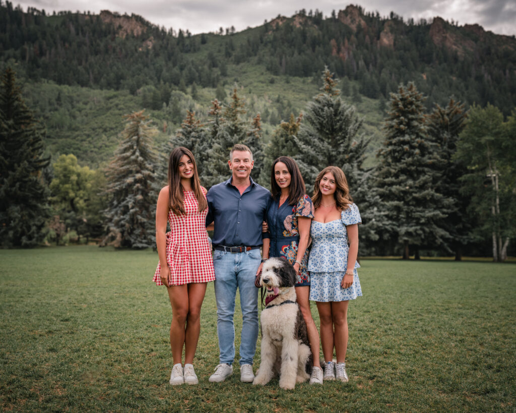 Family standing with their dog in a scenic mountain meadow surrounded by pine trees
