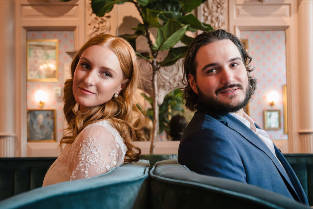 Newlyweds sit back to back in their wedding venue and smile at the camera.