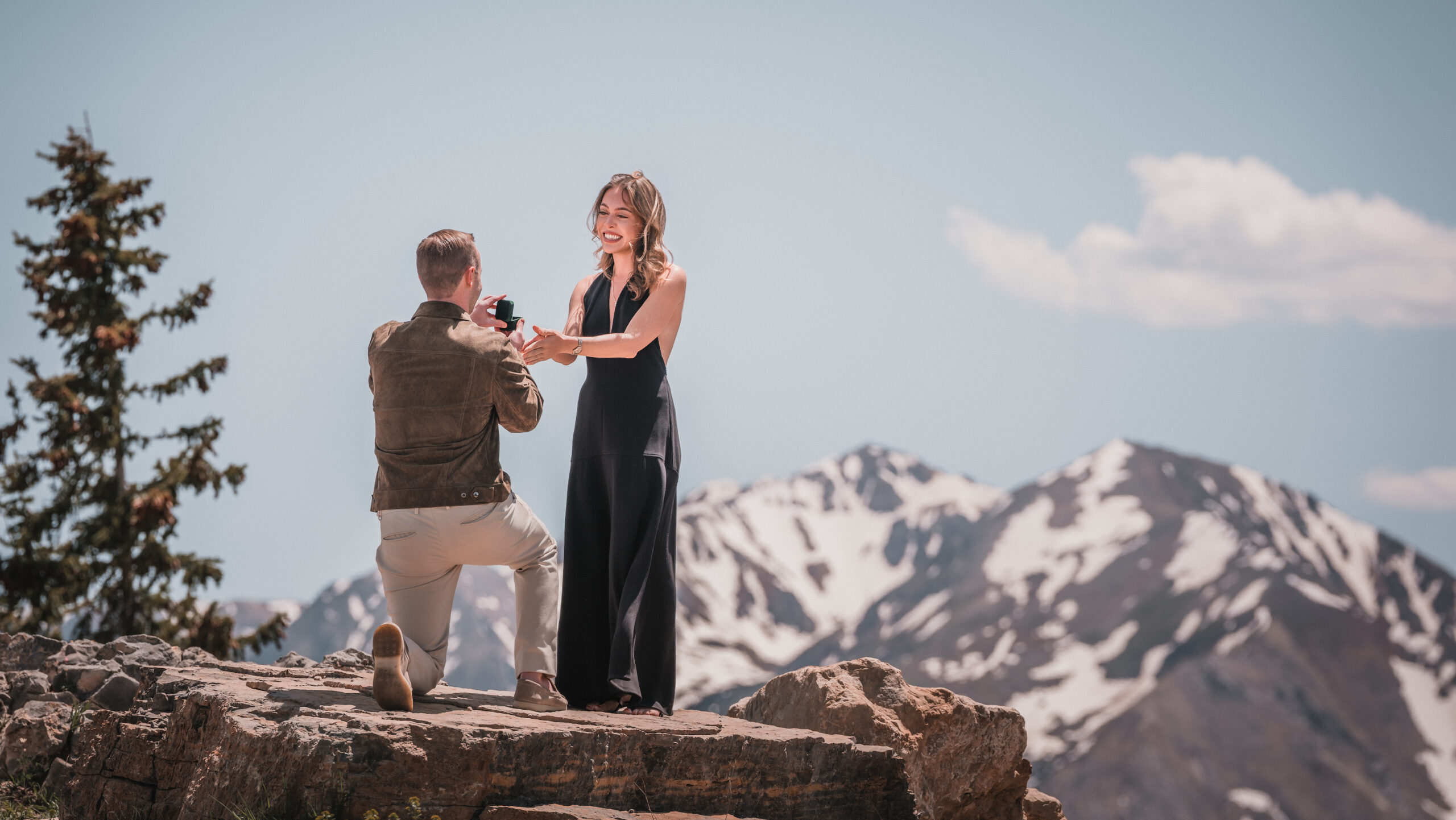Romantic mountain proposal with partner kneeling and holding a ring box on a cliff overlooking the peaks