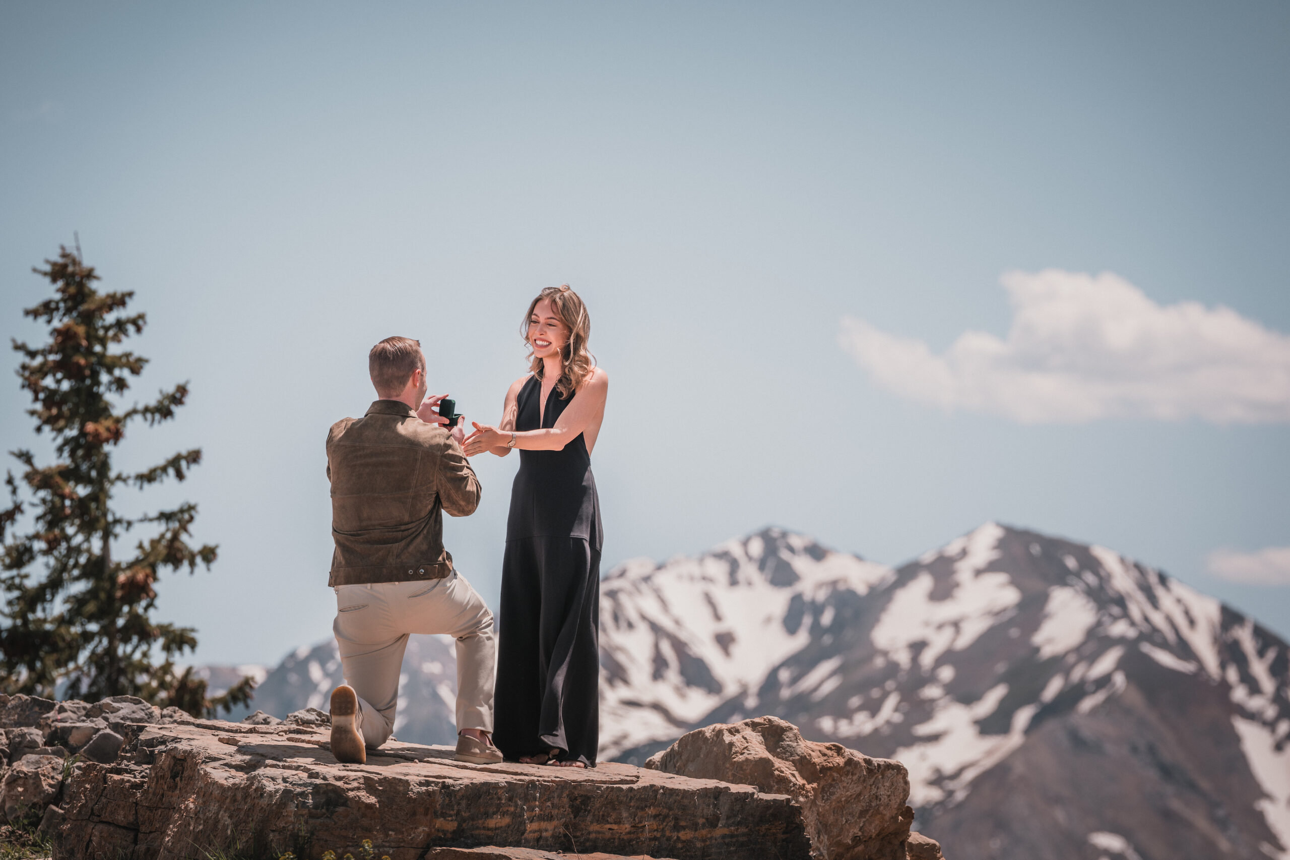 Romantic mountain proposal with partner kneeling and holding a ring box on a cliff overlooking the peaks