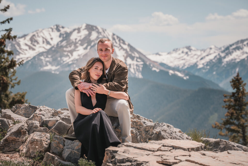Couple sitting closely together on rocky terrain with panoramic mountain views during a wedding engagement photography session.