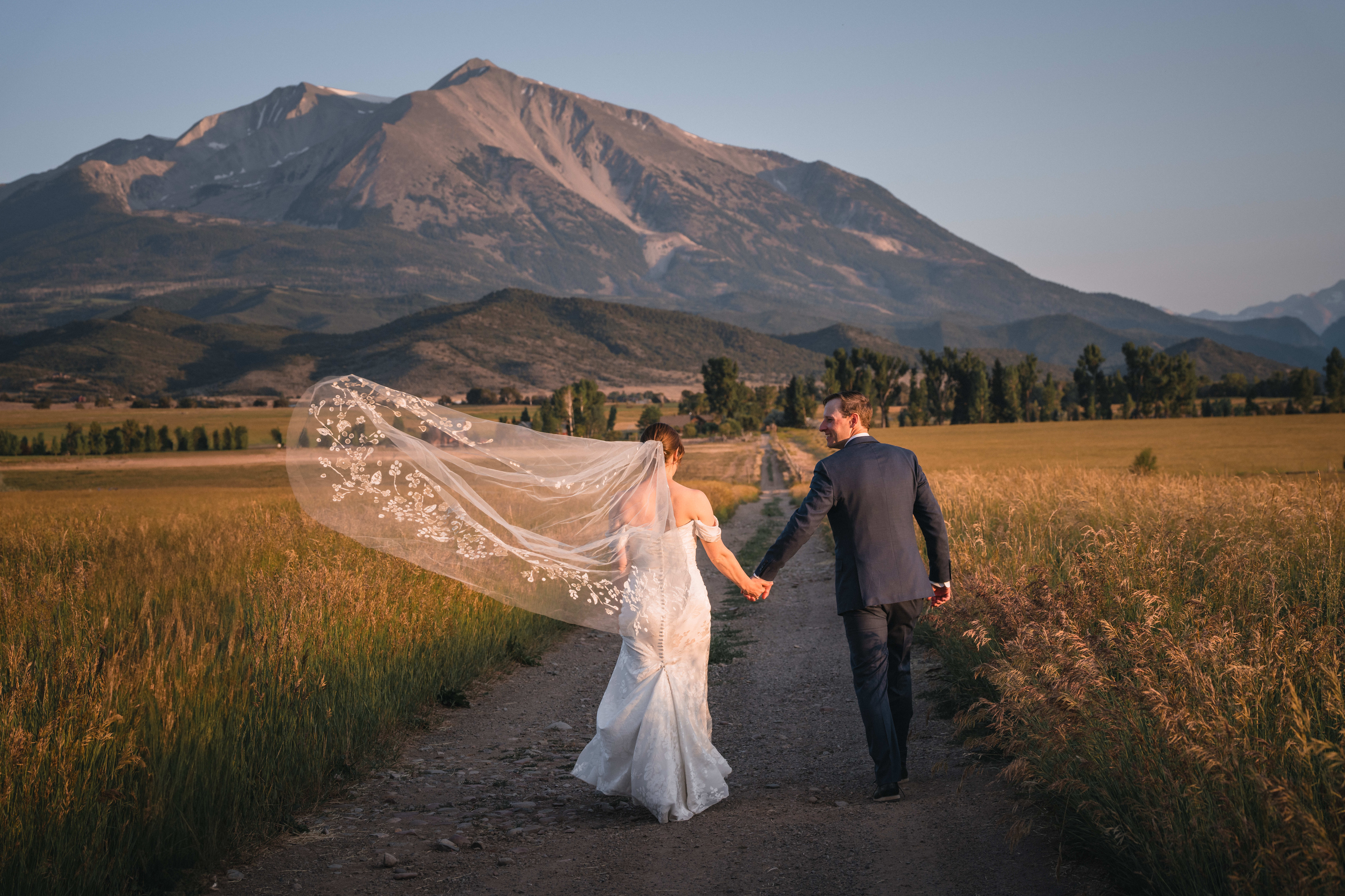 A bride and groom look at each other and smile while the bride's veil blows in the wind. They are walking down a path toward the Aspen mountains.