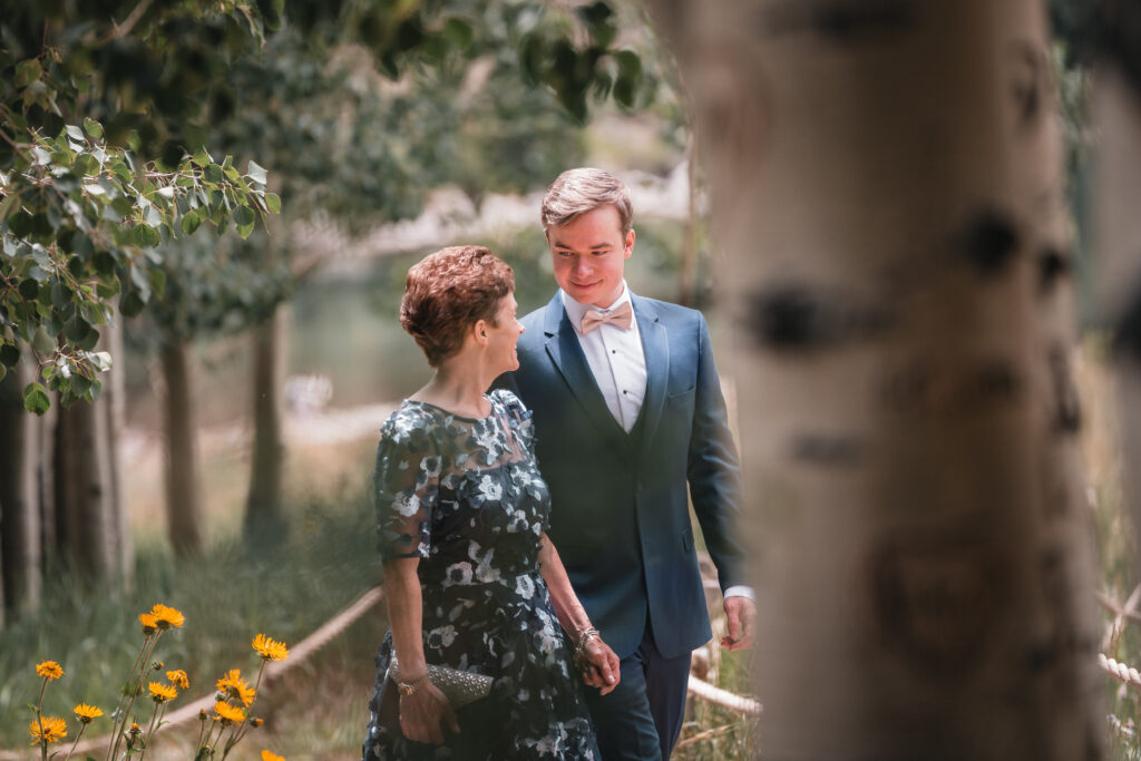 Groom in light blue suit with bow tie walking with mother in floral dress beside wildflowers and aspen trees - Maroon Bells Aspen wedding processional moment