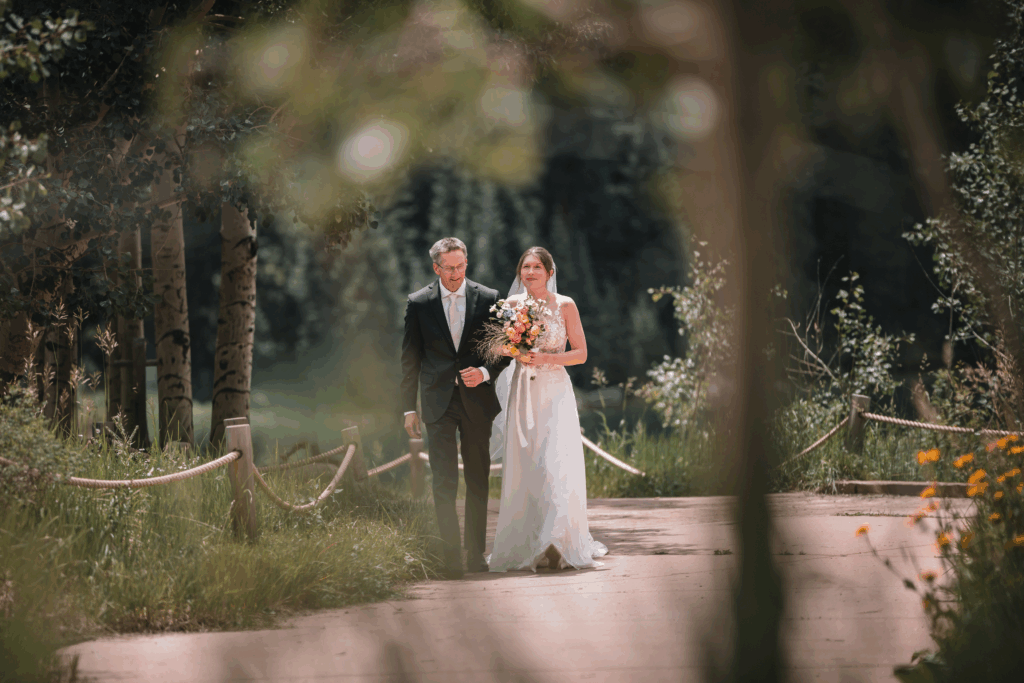 A bride in a white gown holding a colourful bouquet walks down a forest path with a man in a suit, surrounded by lush greenery and sunlight filtering through the trees.