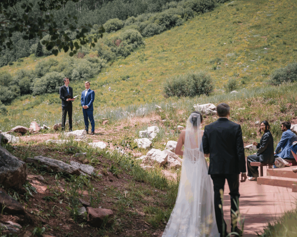 Bride walking down the aisle toward the groom during an outdoor mountain wedding ceremony surrounded by greenery in Colorado.