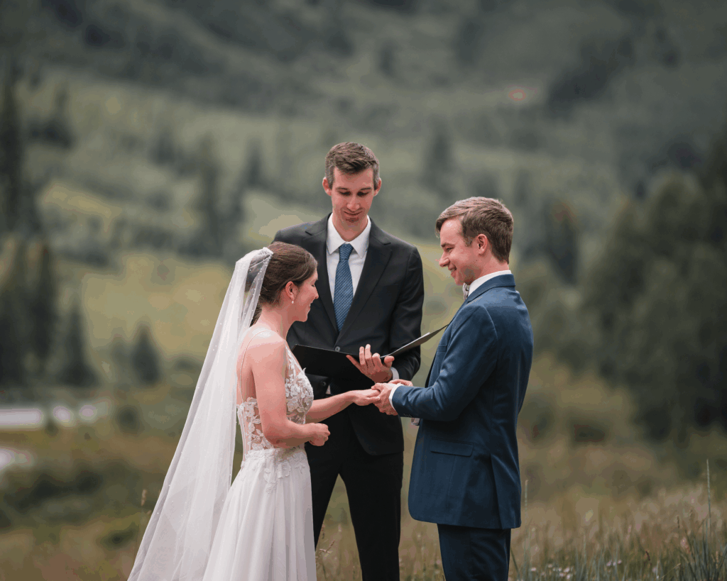 Outdoor mountain wedding ceremony with a bride and groom exchanging rings and an officiant standing between them.