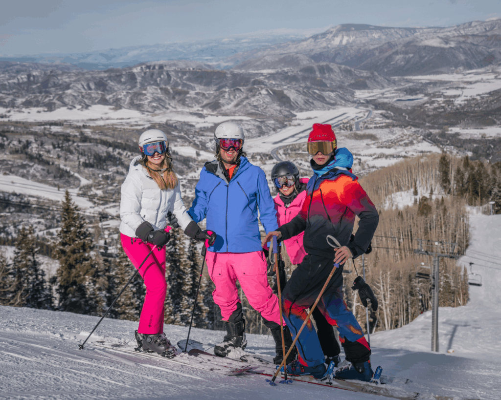 Family in ski gear posing on a snowy peak with panoramic views of the Aspen Mountains in the background.