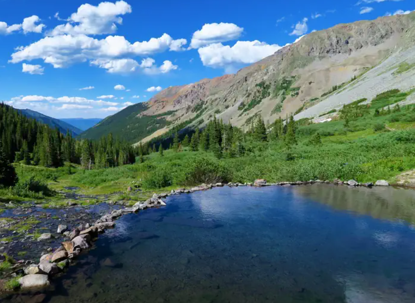 The Maroon Bells’ iconic twin peaks rise above a serene alpine lake, surrounded by pine forests and wildflowers, with their reflection mirrored in the water.