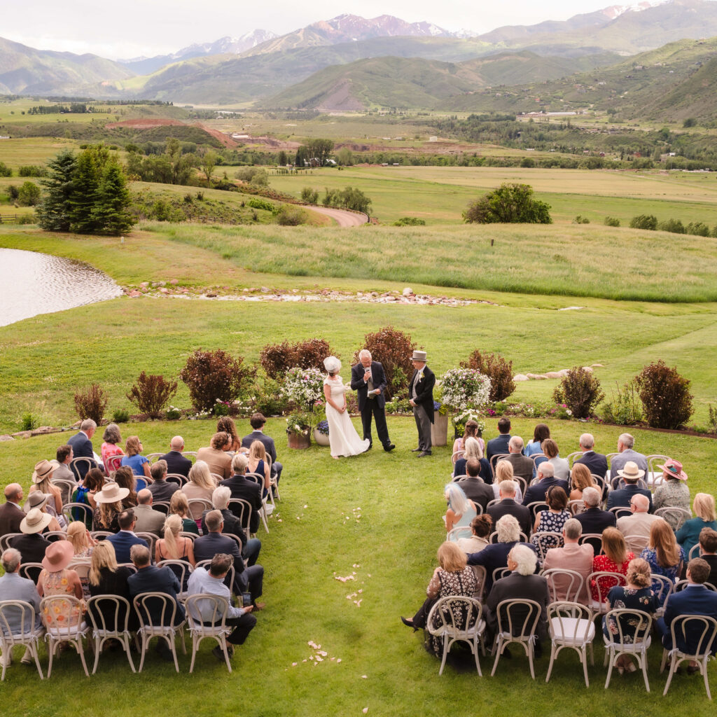 An outdoor wedding ceremony set on a lush green lawn with guests seated in rows facing a couple and officiant. Behind them is a scenic view of rolling hills, a pond, and distant snow-capped mountains under a partly cloudy sky.