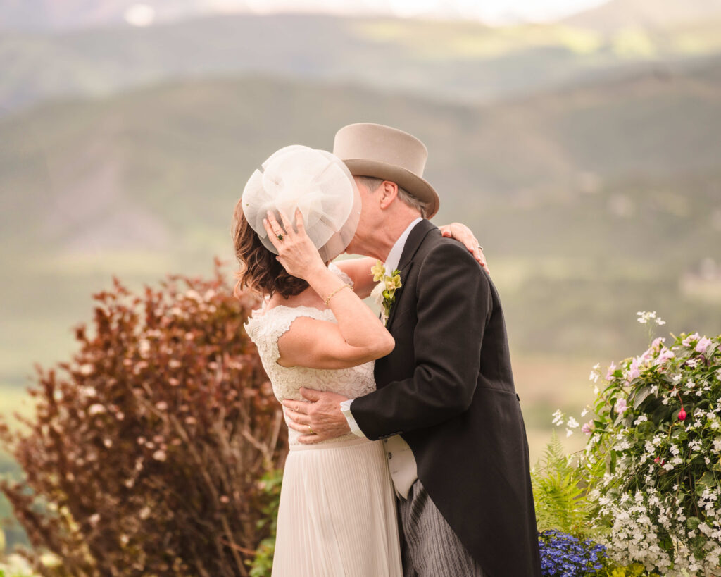 Bride and groom sharing a kiss during their outdoor wedding ceremony, with the bride wearing a white lace wedding dress and veil, and the groom in a dark suit and tan fedora hat, set against a scenic mountain landscape backdrop with floral arrangements visible in the foreground