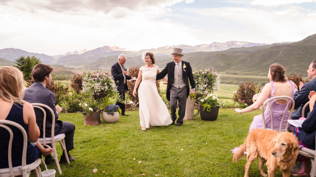 A groom and his new wife walk hand in hand down the aisle, smiling as the Aspen mountains rise behind them.