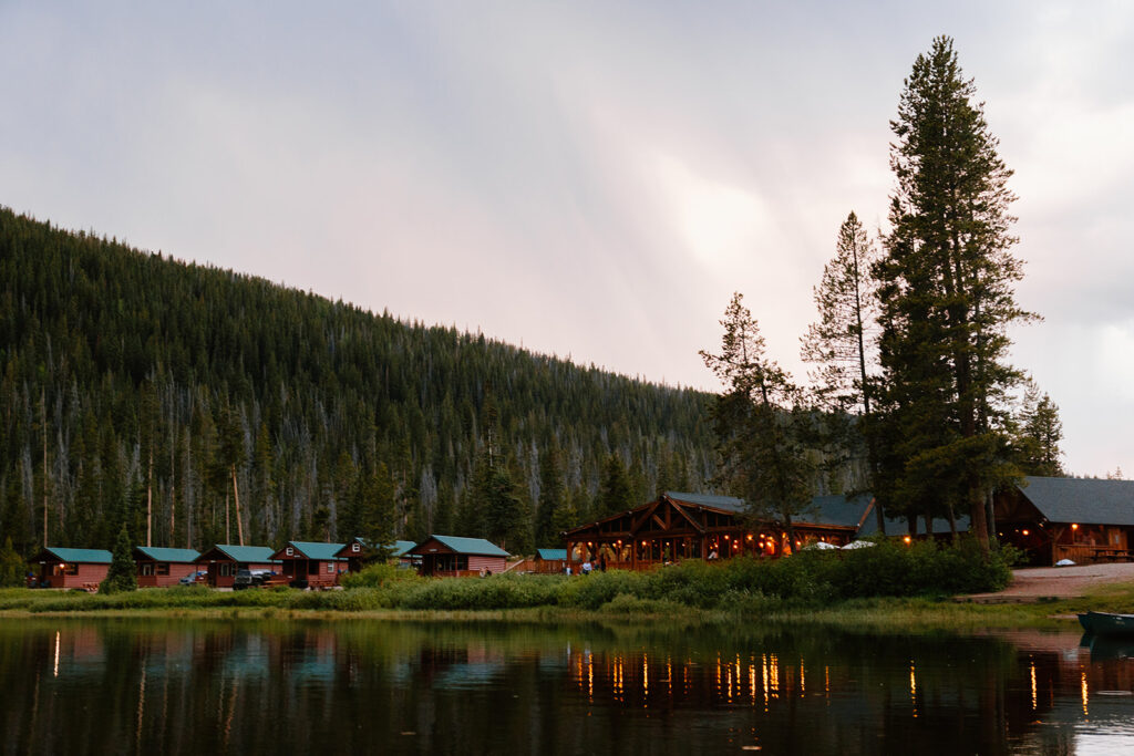 Scenic lakeside wedding venue at Piney River Ranch in Vail, Colorado, featuring rustic log cabins, a lodge with warm lights, and mountain forest views reflected on the water.