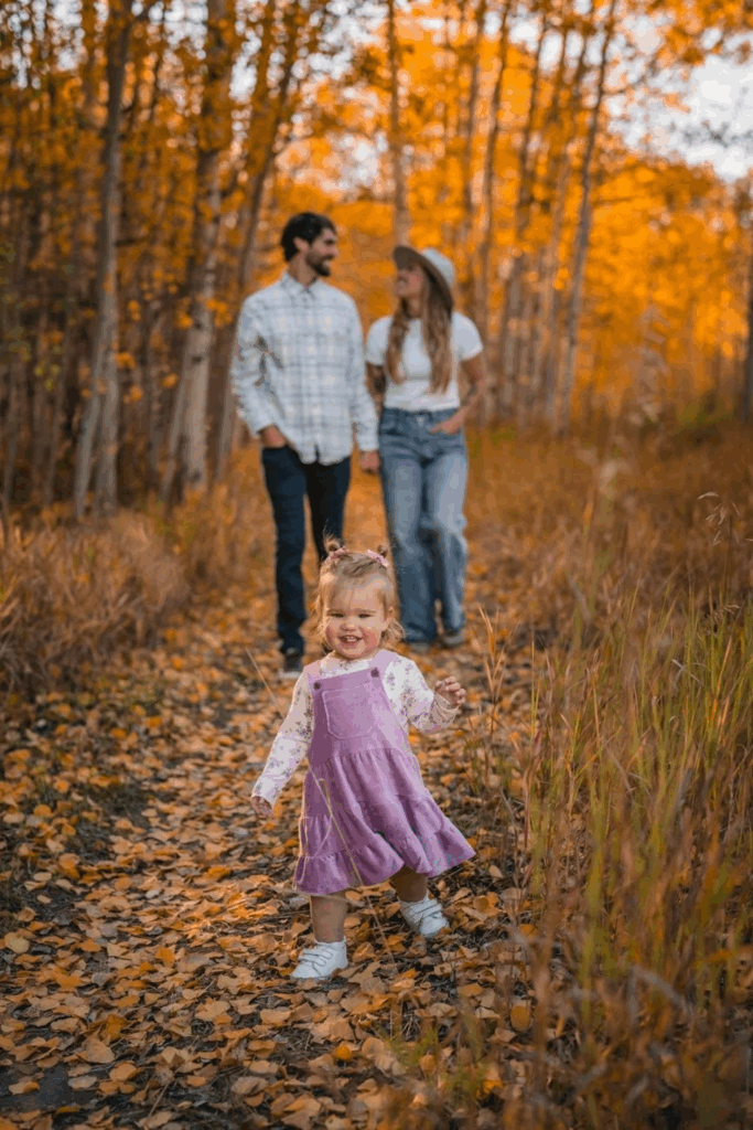 Husband and wife walk in the fall leaves with trees surrounding them as their baby girl runs ahead of them.