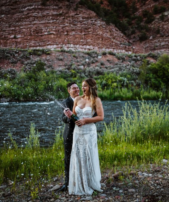 The bride, holding champagne, and the groom laugh together in front of a sparkling stream at Spring Creek Ranch, surrounded by lush greenery