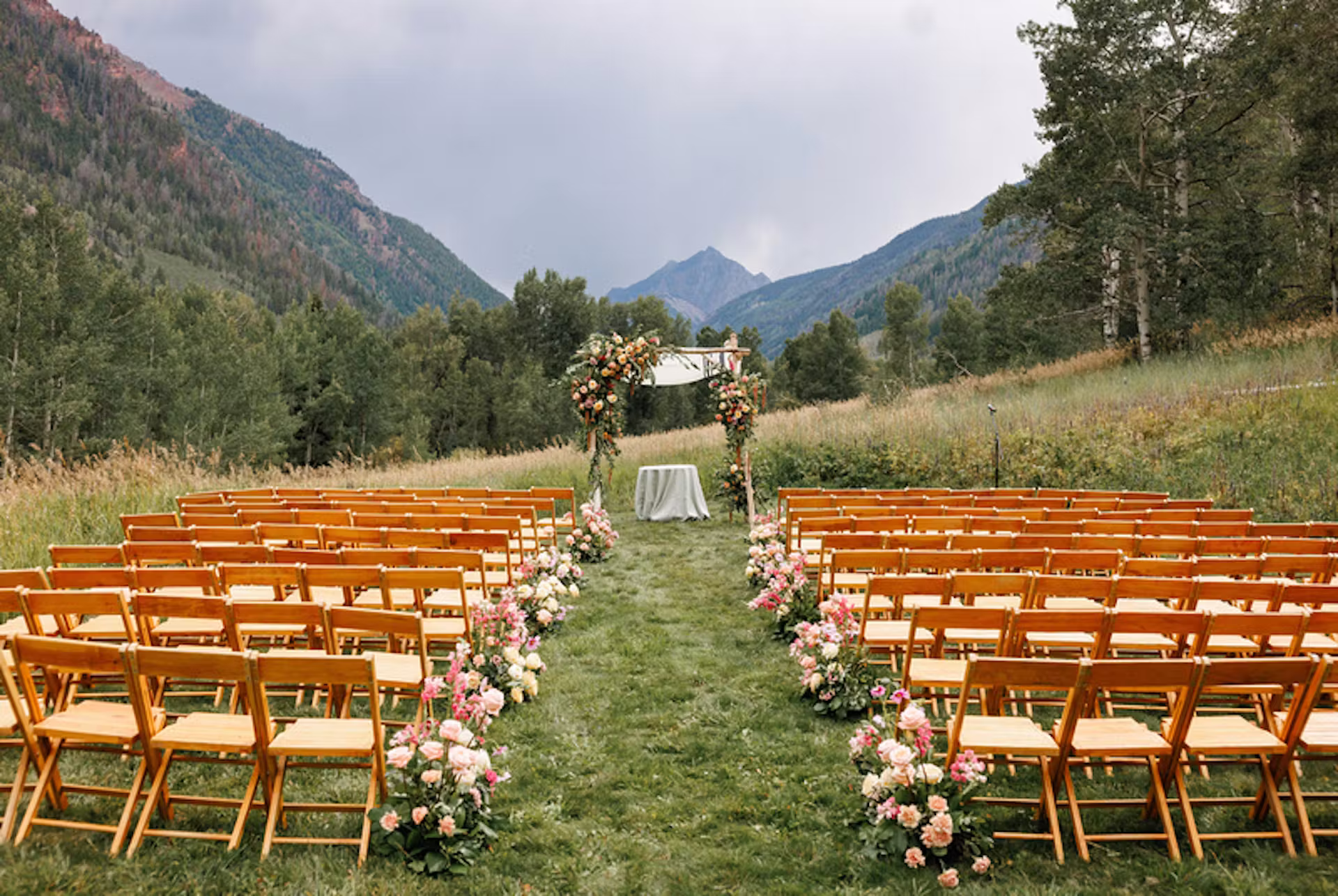 Elegant outdoor mountain wedding ceremony with wooden chairs, lush flower arrangements lining the aisle, and a floral arch set against panoramic alpine views.